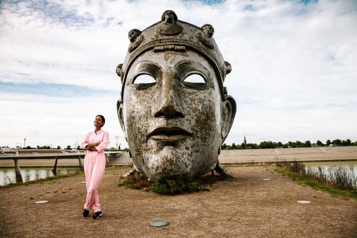 Deborah in front of face of Nijmegen. On the city island of Veur-Lent you will find a Roman mask, made by artist Andreas Hetfel. It is also called the face of Nijmegen and you can hardly miss it, once you enter the island.  One of the best things to do in Nijmegen if you want to take fun pictures.